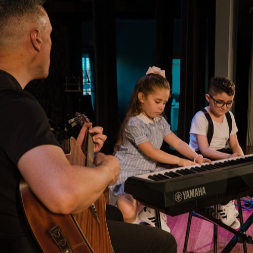 Professor tocando violão ao lado de duas crianças no teclado durante a aula