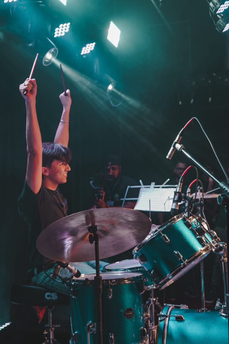 Aluno da Da Capo Musical sorrindo com as baquetas para o alto, na bateria, durante apresentação no palco