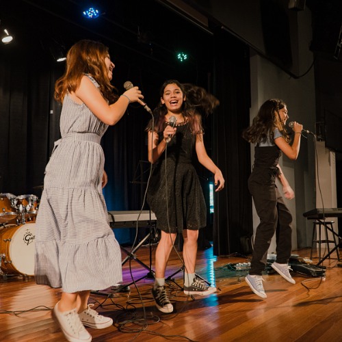 Três alunas cantando juntas ao microfone durante apresentação