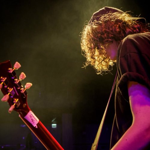 Aluno tocando guitarra no palco, com o cabelo iluminado pelo refletor